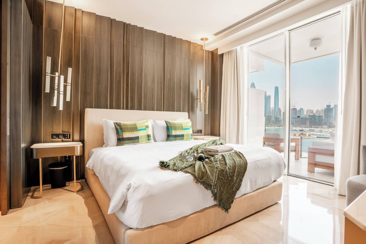 Una habitación de hotel moderna con una cama grande, una mesita de noche, vistas de la ciudad a través de una puerta de vidrio y una pared de acento de madera.