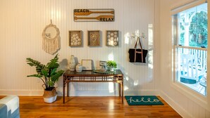 Una habitación con piso de madera, una mesa con un tapete que dice 'hello', un letrero que dice 'relajación en la playa', un colgador de macramé y una ventana con vista a una terraza.