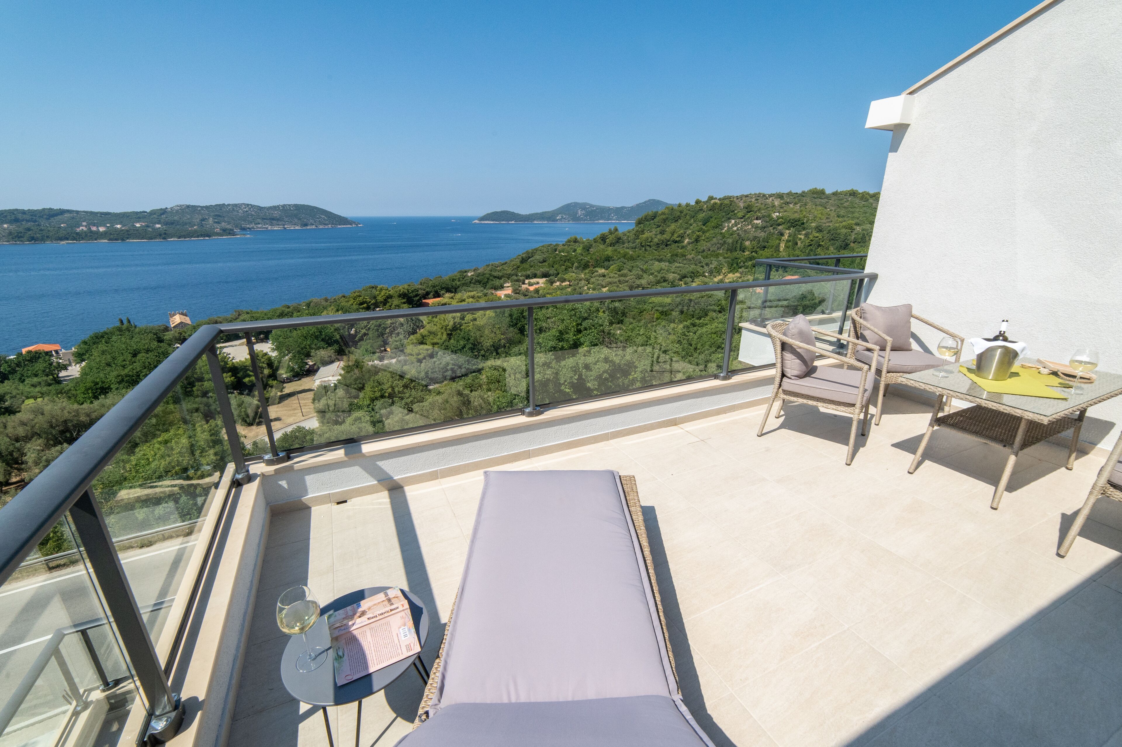 A balcony with a view of the sea, a lounge chair, a table with a bottle and glasses, and a small table with a menu.