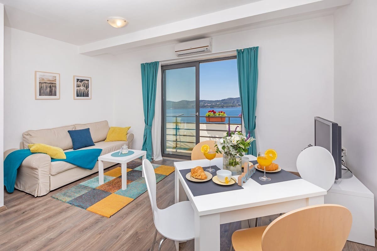 Uma sala de estar moderna com sofá, mesa de centro e mesa de jantar posta para o café da manhã. Uma varanda com vista para o mar é visível pela janela.