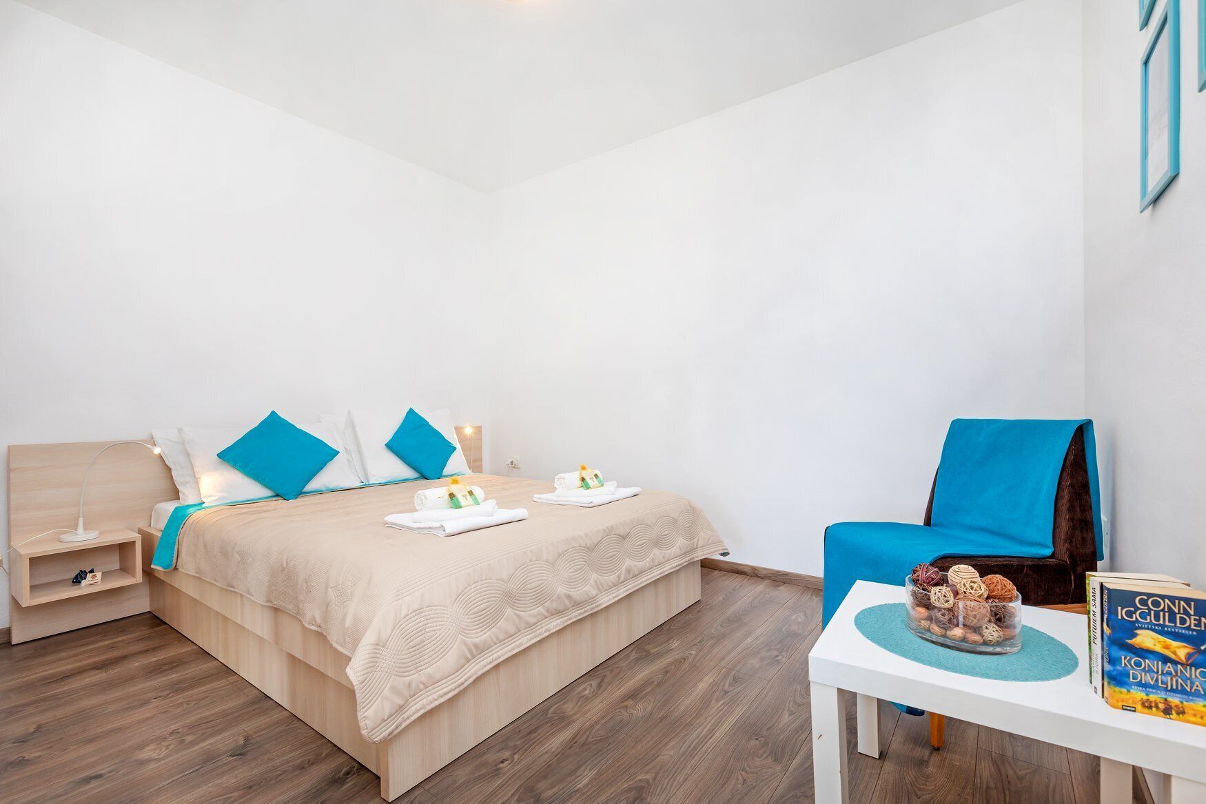 A modern hotel room with a bed, a chair, a nightstand, and a table with a book and decorative items.