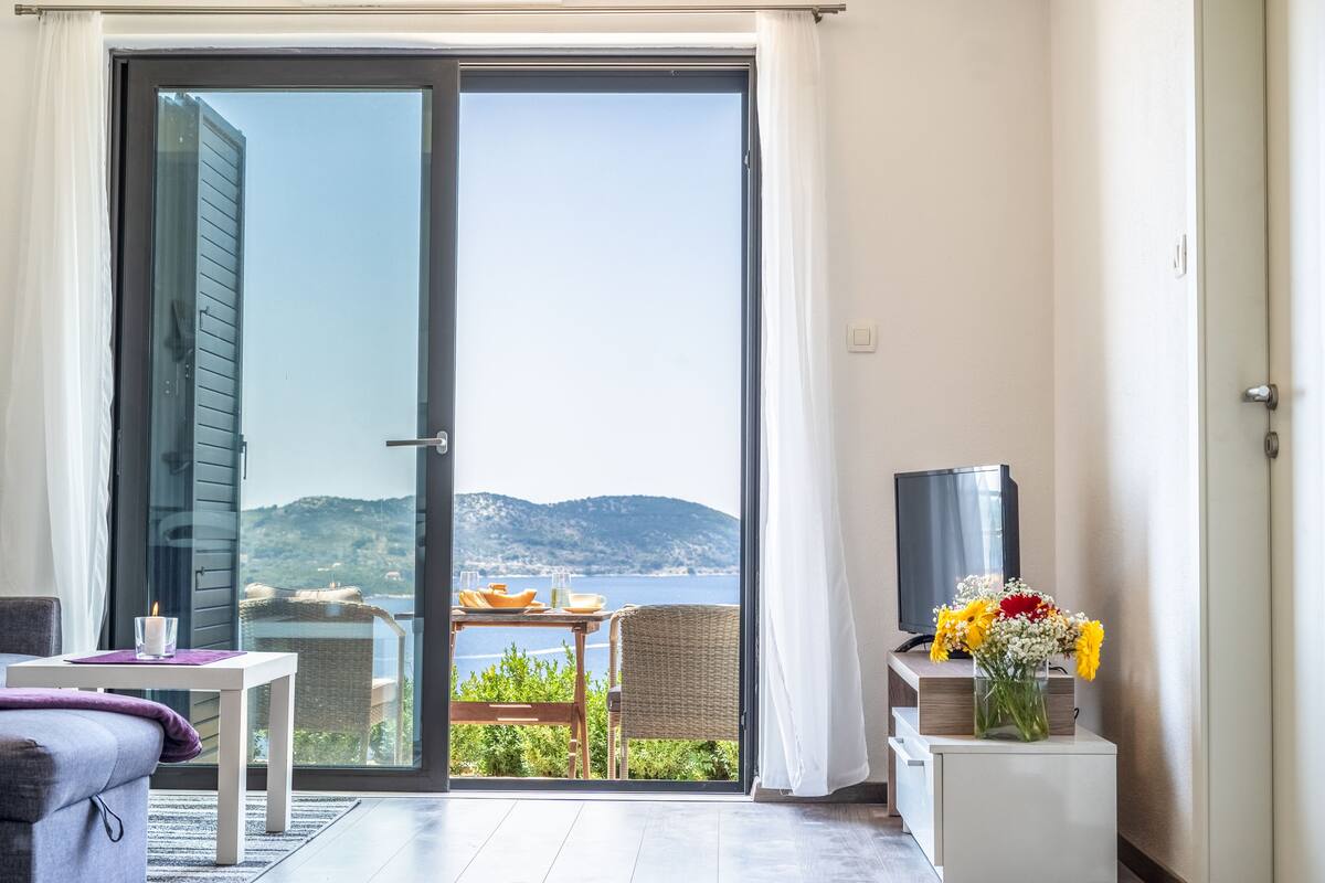 Quarto com porta de vidro deslizante que dá para uma varanda com vista para o mar e colinas. Há uma mesa com vela, copos e um prato. Uma TV está sobre um suporte ao lado da porta.