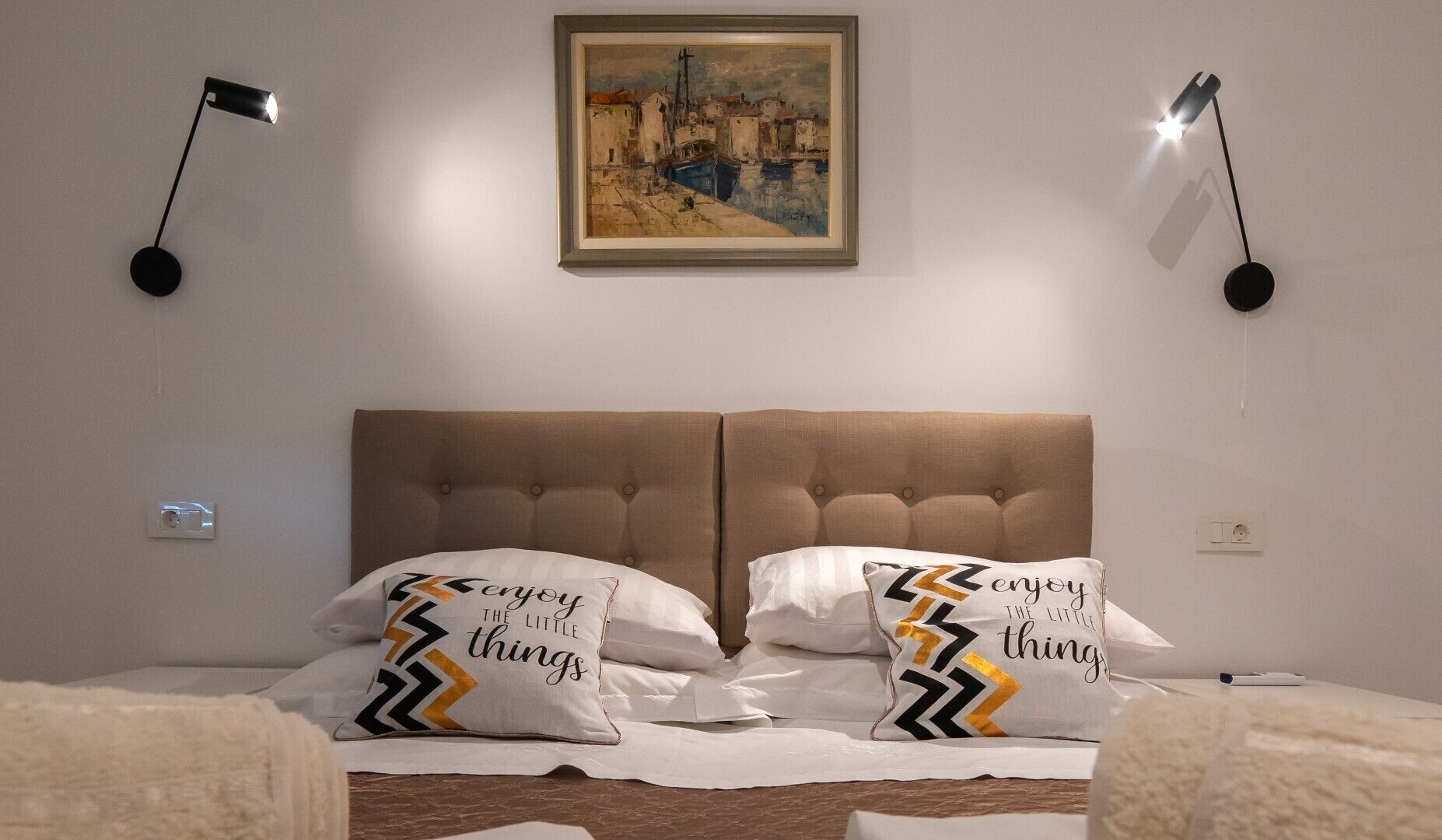 A hotel room with a large bed, two wall-mounted reading lamps, a framed painting above the bed, and decorative pillows with a chevron pattern.