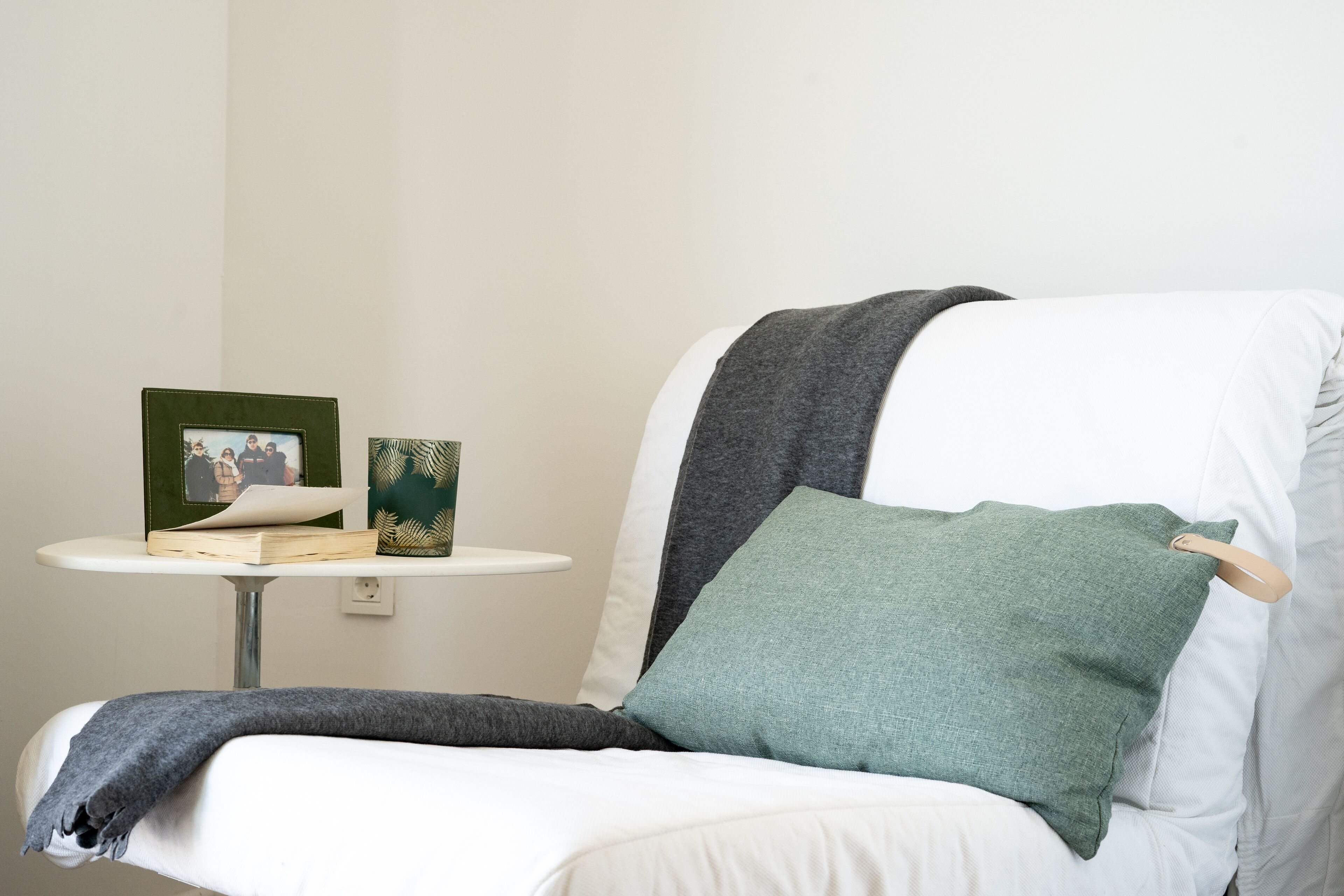 Una silla con un cojín verde, una manta gris y una mesita con un marco de fotos y un adorno decorativo verde.