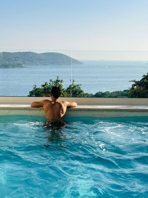 En person slapper af i en pool med udsigt til havet og bakker.