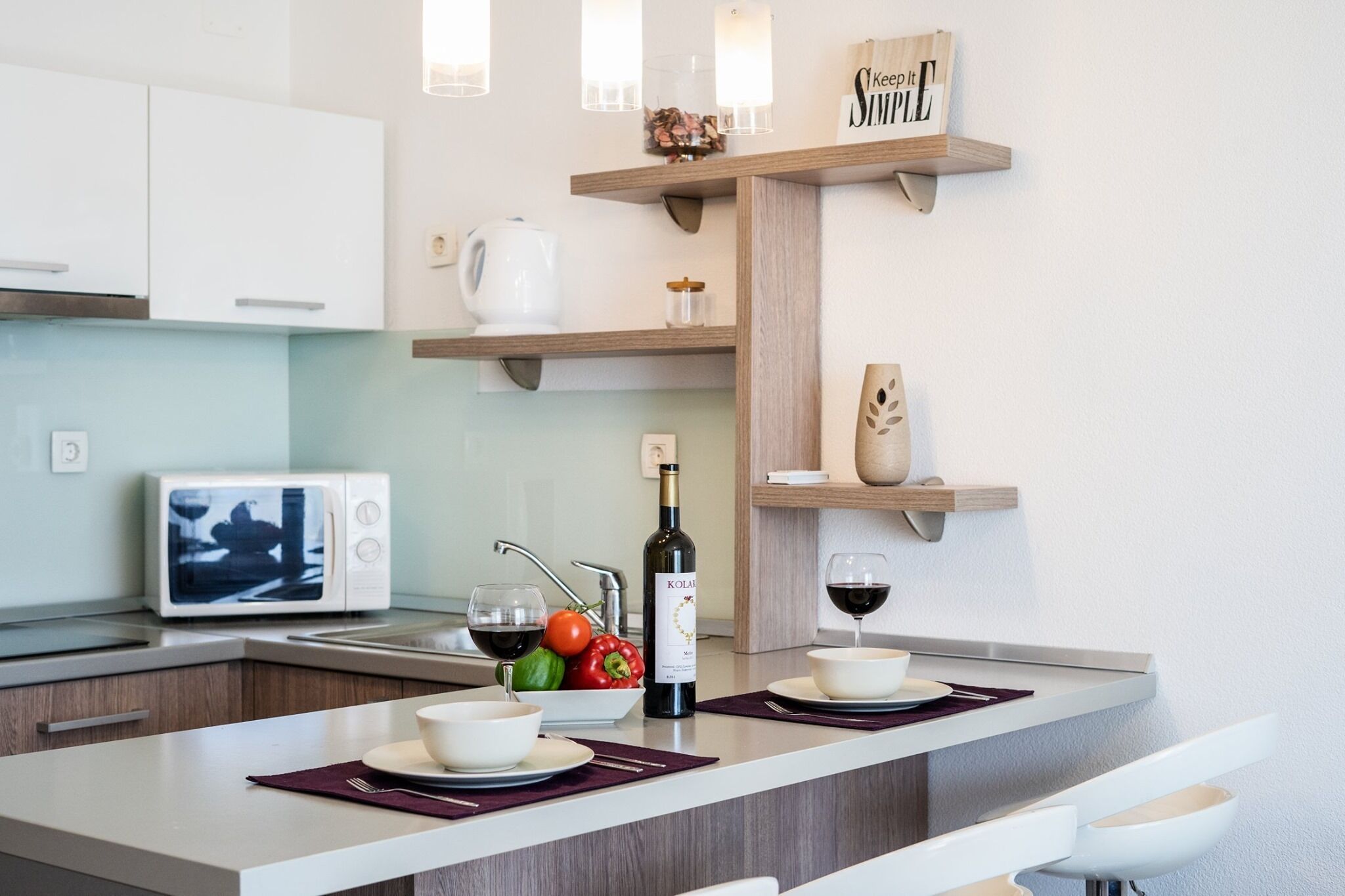 A modern kitchen with white cabinets, a built-in microwave, a wine bottle, and a fruit bowl.