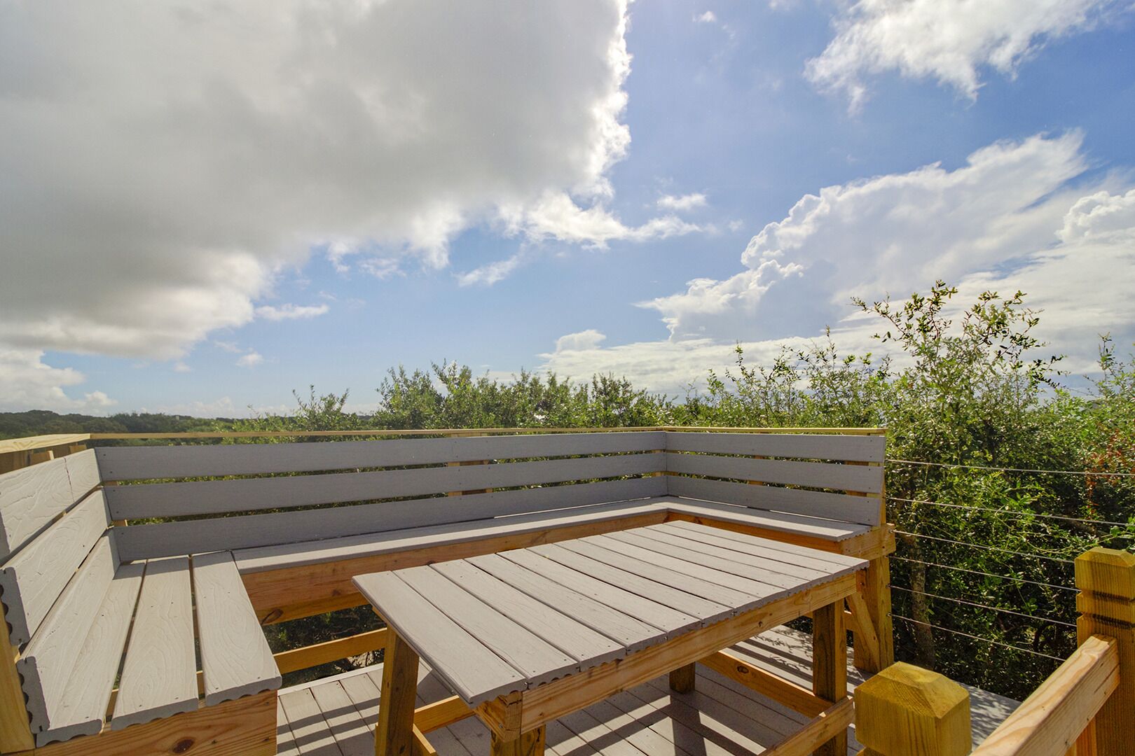 A wooden deck with a bench and a table, surrounded by greenery and a cloudy sky.
