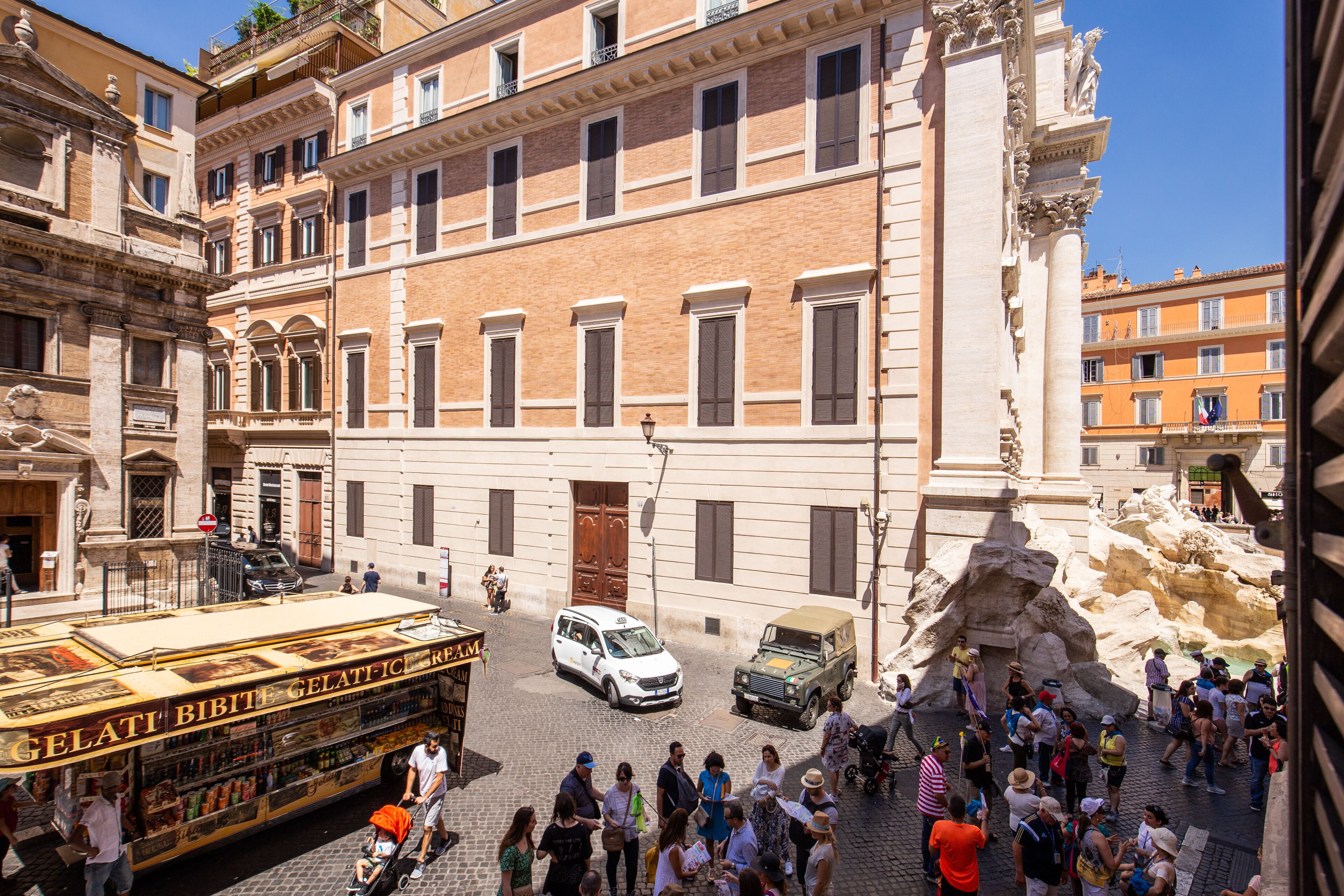 A crowded street with tourists, a gelateria, and a fountain.
