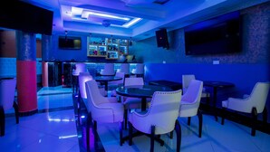 A bar area with white upholstered chairs, a bar counter, and a television mounted on the wall. - Hotel San Marino (Kitui)