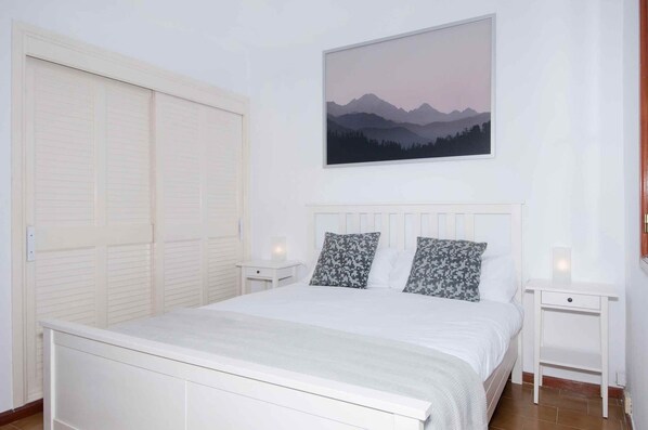 A bedroom with a bed, two nightstands, a white headboard, a large window, and a mountain landscape painting.