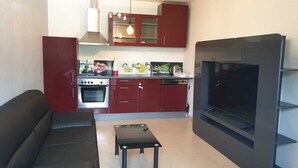 Un salon moderne avec un canapé en cuir noir, une petite table basse noire, un téléviseur à écran plat et un coin cuisine doté de meubles rouges et d’une hotte de cuisine en acier inoxydable.