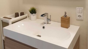 Une salle de bain moderne avec un grand lavabo blanc, un miroir, une petite plante en pot, un porte-savon en bois et un porte-serviettes.
