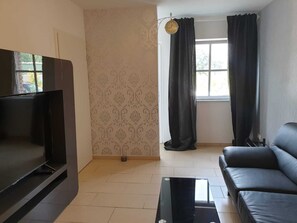 A living room with a flat-screen TV, a black leather sofa, a glass coffee table, patterned wallpaper, and a window with curtains.