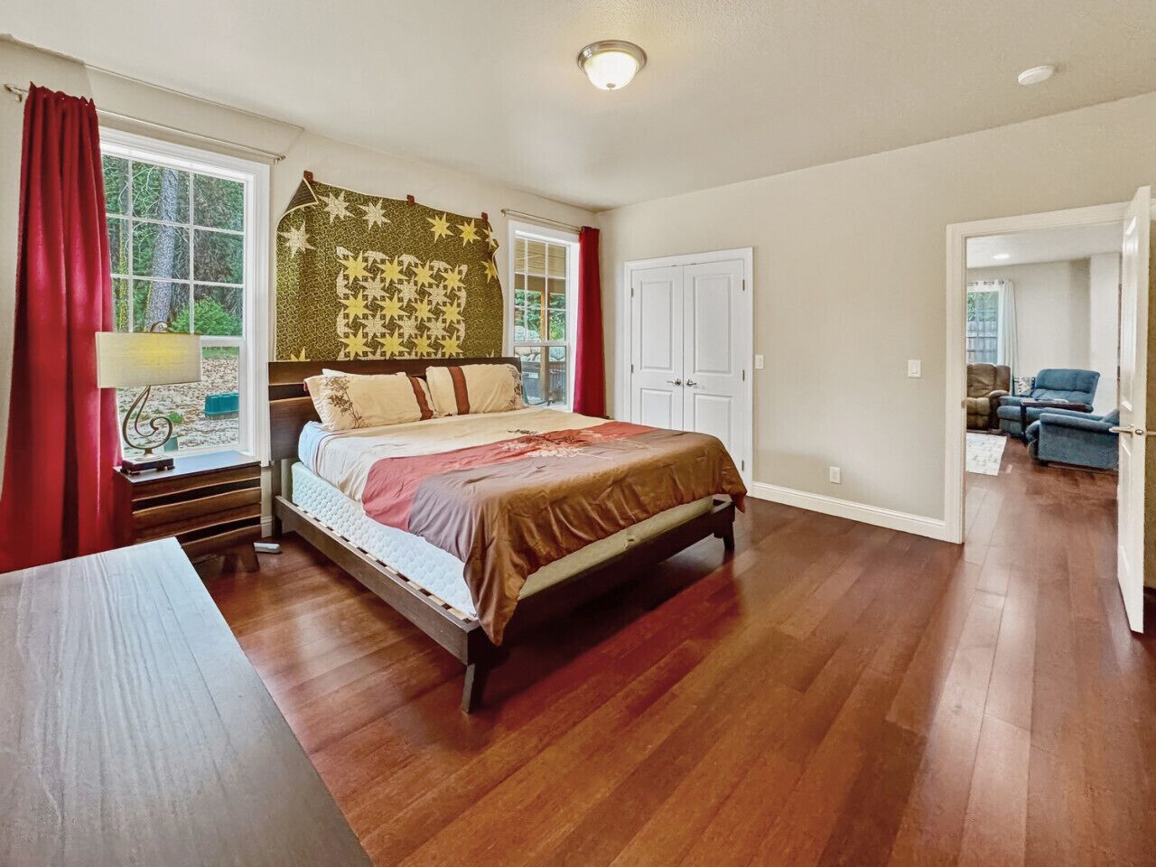 A bedroom with a wooden floor, a bed with a brown bedspread, a window with red curtains, and a door leading to another room.