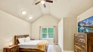 Une chambre à coucher avec un lit en bois, une table de chevet, une commode, un ventilateur de plafond et une fenêtre avec des rideaux.