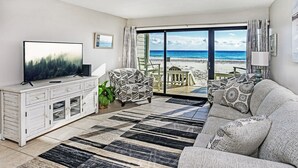 A living room with a large flat-screen TV, a sofa, a patterned armchair, and a view of the beach through a sliding glass door.
