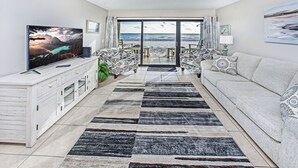 A living room with a large flat-screen TV, a sofa, patterned rugs, and a view of the beach through glass doors.