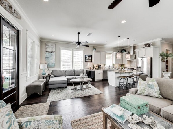 A spacious living room with a sofa, coffee table, and ottomans. There is a kitchen area with a refrigerator, stove, and bar stools.