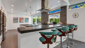 A modern kitchen with a central island, bar stools, and a large window providing a view of the outdoors. - Iconic Twin Palms - Classic Upscale Midcentury! (Palm Springs)