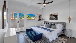 A bedroom with a bed, a ceiling fan, a bench, and a view of palm trees through the door. - Iconic Twin Palms - Classic Upscale Midcentury! (Palm Springs)