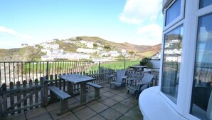 A patio with wooden tables and chairs, a view of a hillside with white buildings, and a clear sky.