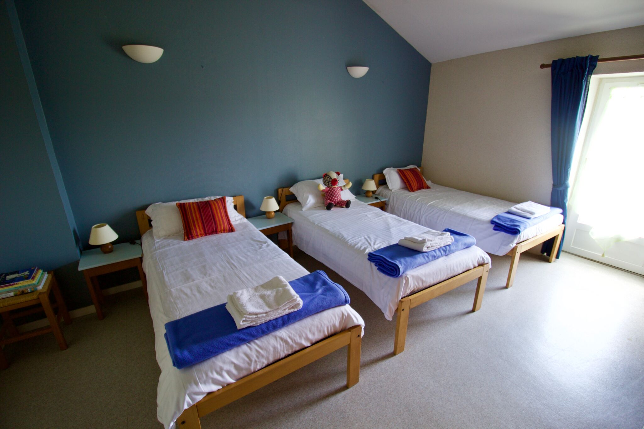 A hotel room with three single beds, each with a pillow and a folded towel. There are bedside tables with lamps and a small table with books. The room has a blue wall and a window with curtains.