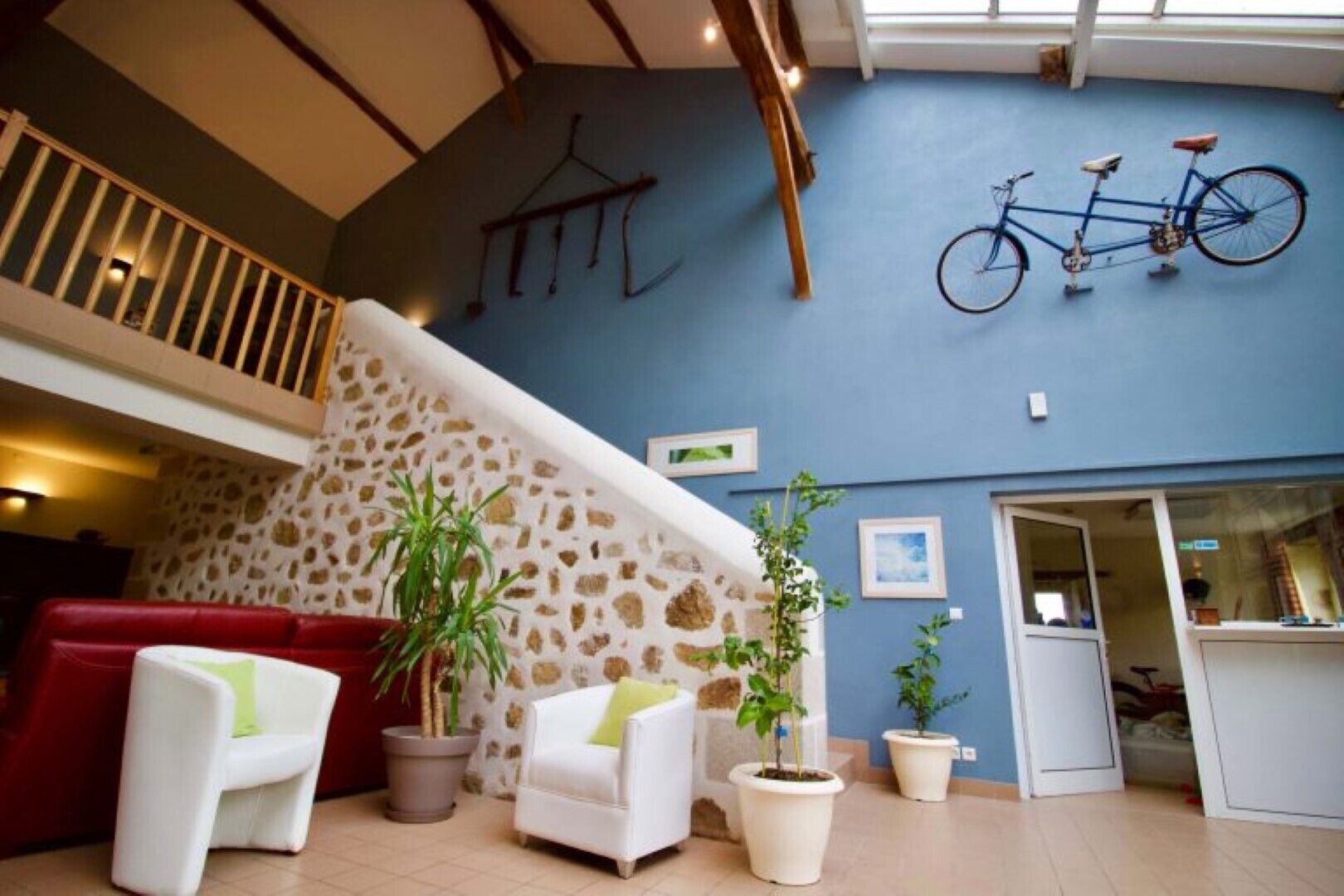A spacious lobby with a stone wall, a staircase, a red couch, white armchairs, potted plants, and a bicycle mounted on the wall.