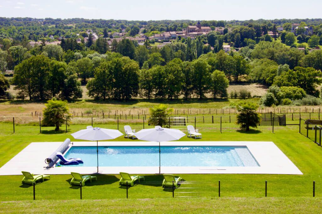 A rectangular swimming pool with a white deck, surrounded by green lawn chairs and umbrellas, set against a backdrop of a rural landscape with trees and houses.