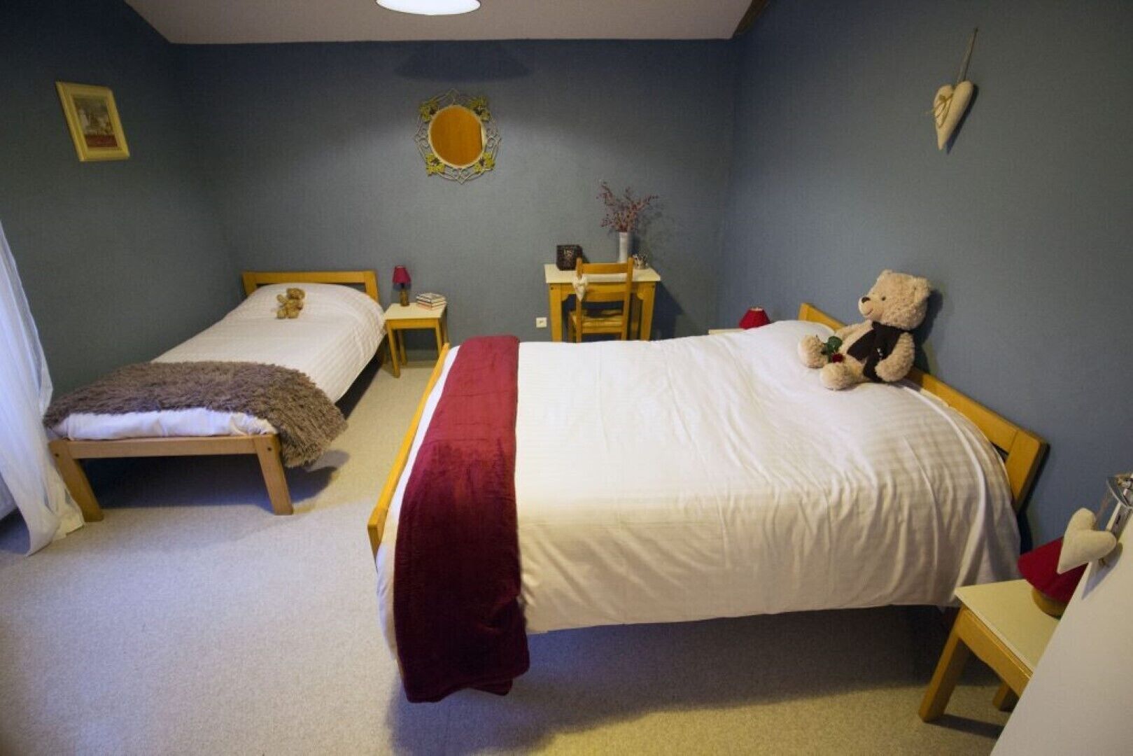 Ein Schlafzimmer mit zwei Einzelbetten, einem Holfnachtkästchen, einem Nachttisch, einer roten Tagesdecke und einem Teddybären auf einem Bett.