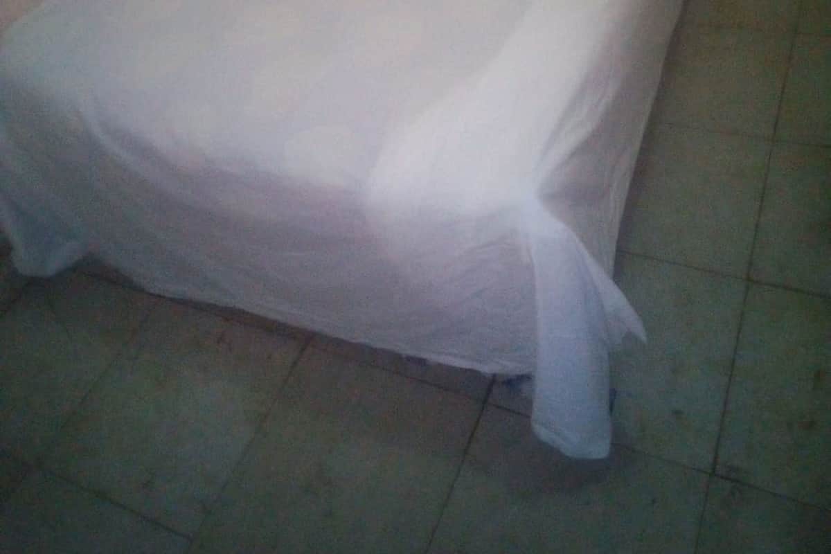 Ein kleines Zimmer mit einem Einzelbett, einer schlichten weißen Bettdecke und einem einfachen, schlichten Kopfteil. Der Boden ist mit Fliesen belegt und die Wände sind schlicht gehalten, mit wenig Dekoration.