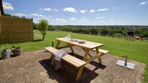 Una mesa de picnic de madera al aire libre con bancos, rodeada de grava, con un campo verde y un cielo azul despejado de fondo.