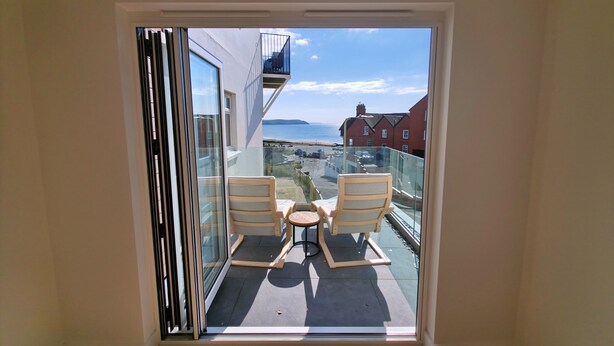 A balcony with two chairs and a small table, overlooking a coastal area with buildings and a view of the sea. - Great dog friendly 2 bed apartment close to village and beach. (Woolacombe)
