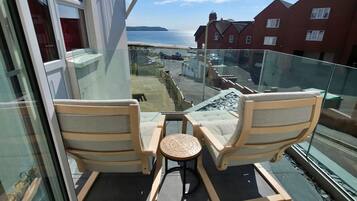 A balcony with two wooden chairs and a small table, overlooking a residential area and a beach.