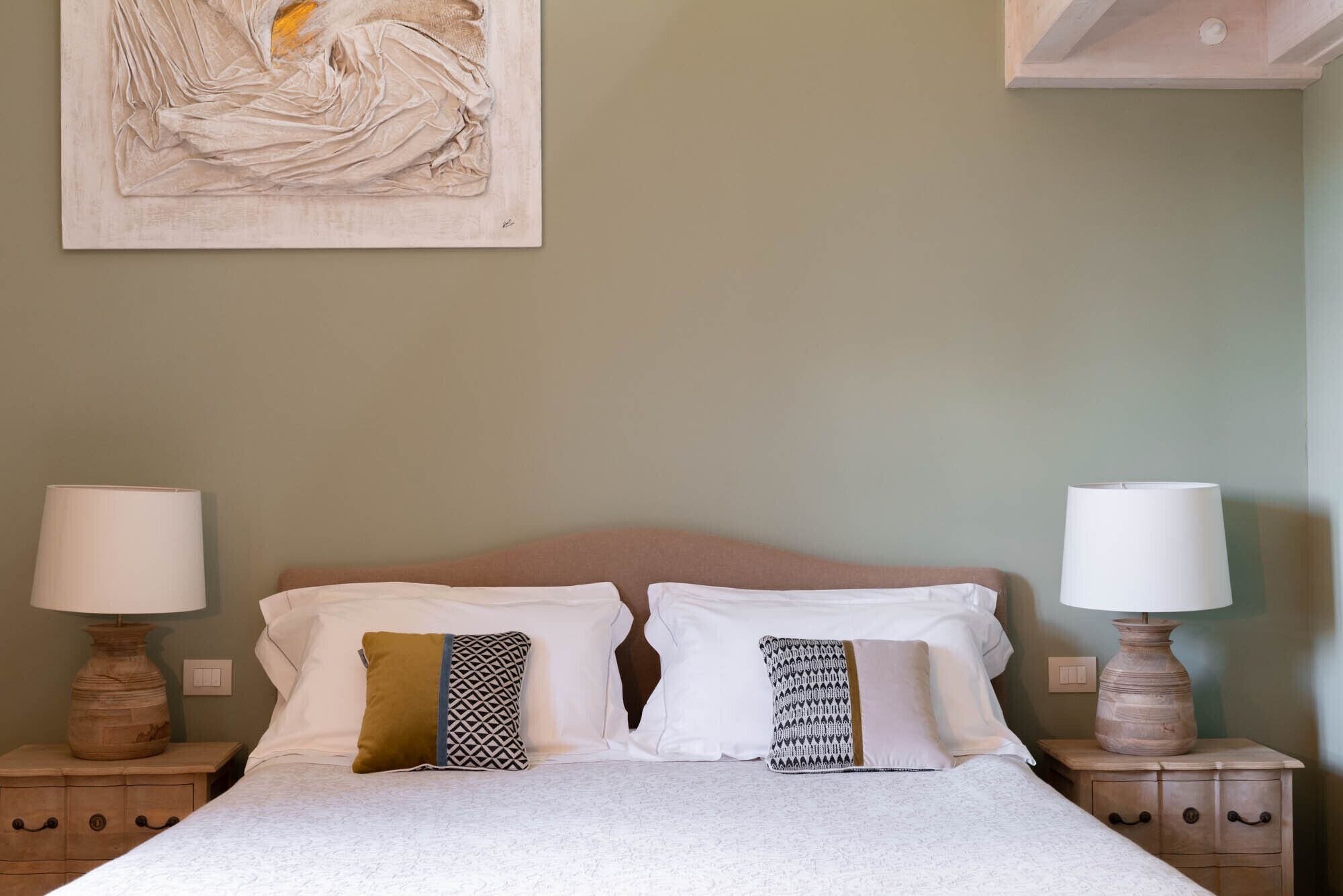A neatly made bed with two pillows, two bedside tables with lamps, and a painting on the wall.