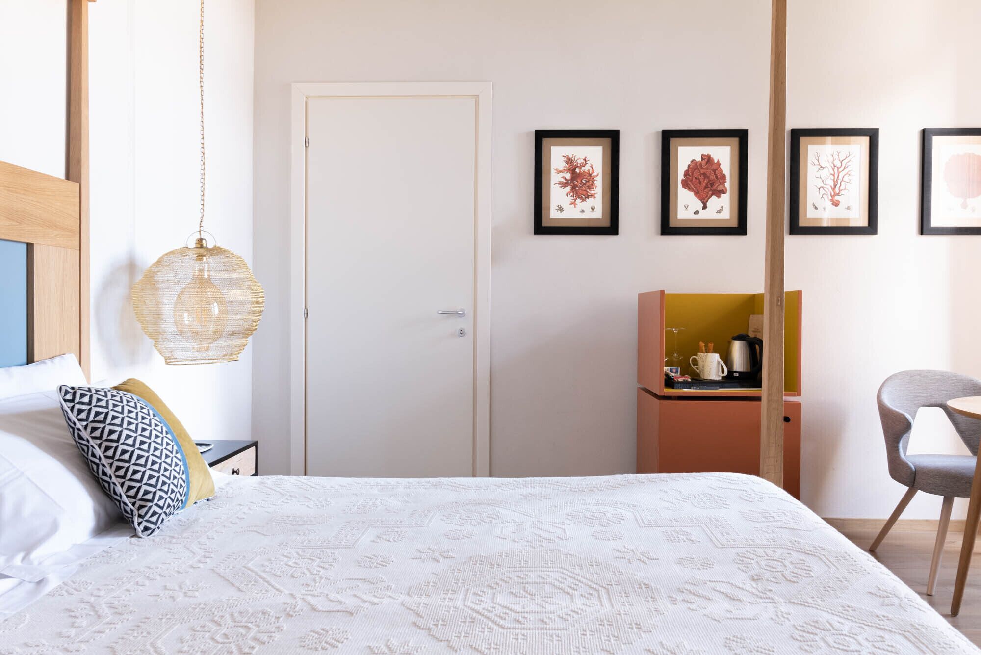 Una camera da letto con un letto, un comodino, una lampada a sospensione, un armadio, un tavolino con un bollitore e opere d'arte incorniciate al muro.
