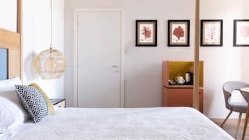 A bedroom with a bed, a bedside table, a hanging lamp, a wardrobe, a small table with a kettle, and framed artwork on the wall.