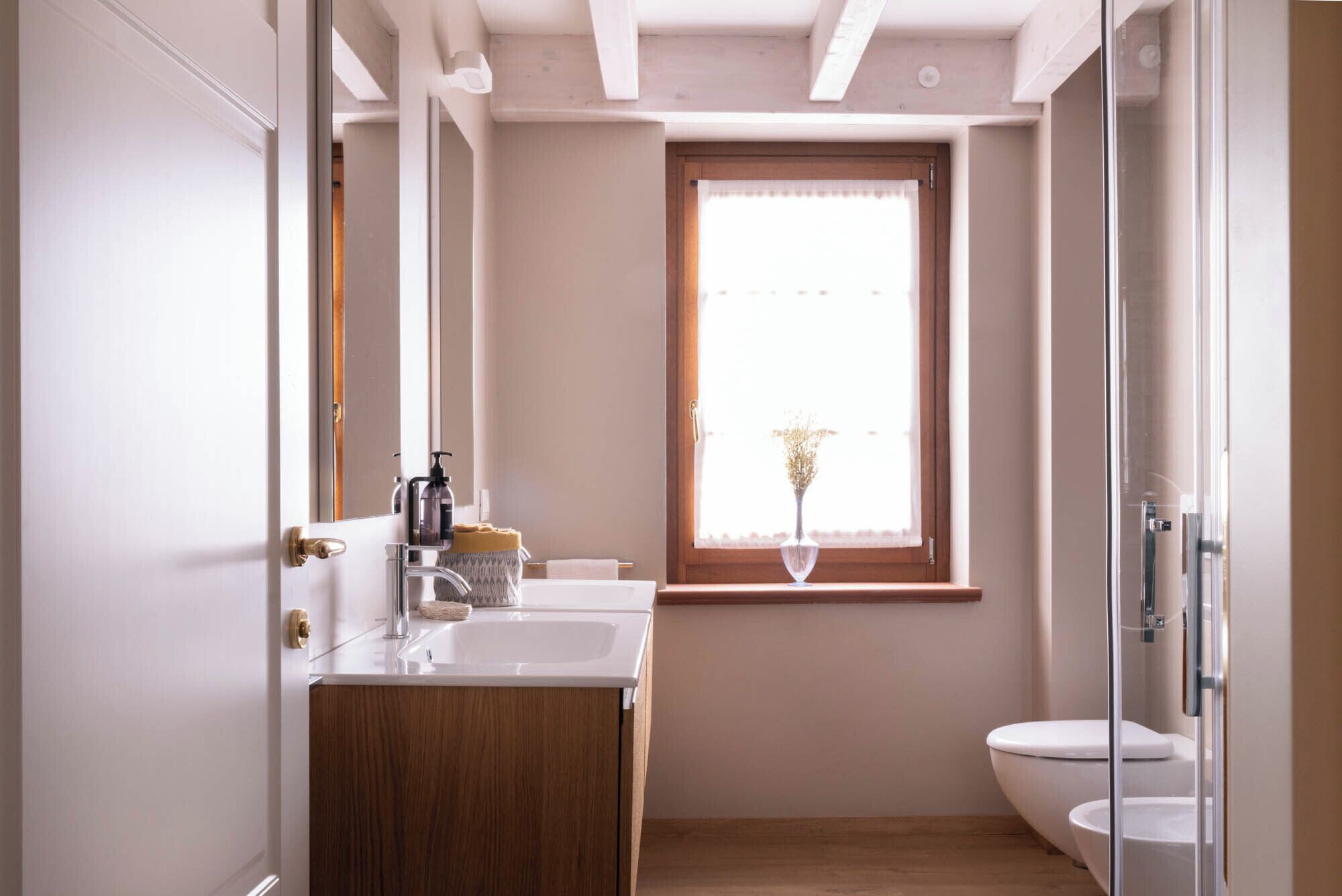 A bathroom with a wooden vanity, a white sink, a glass shower enclosure, and a window with a view of the outdoors.
