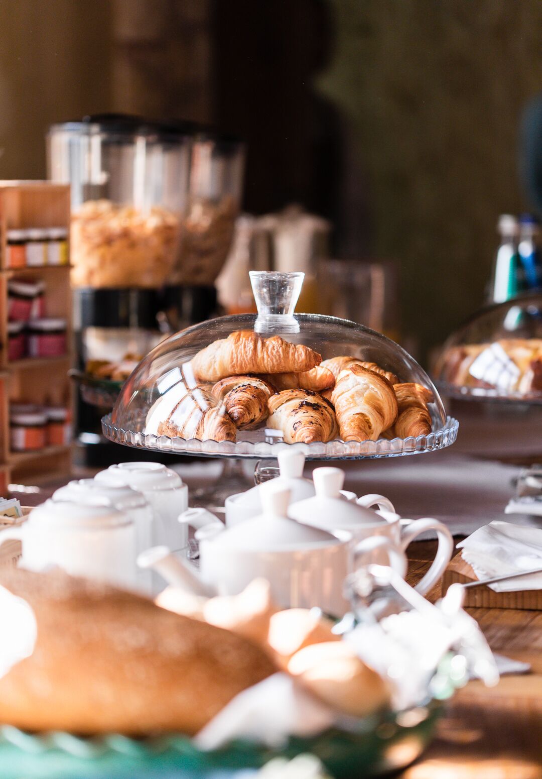 A breakfast buffet with a variety of pastries and bread, including croissants and baguettes, displayed on a glass-covered stand.