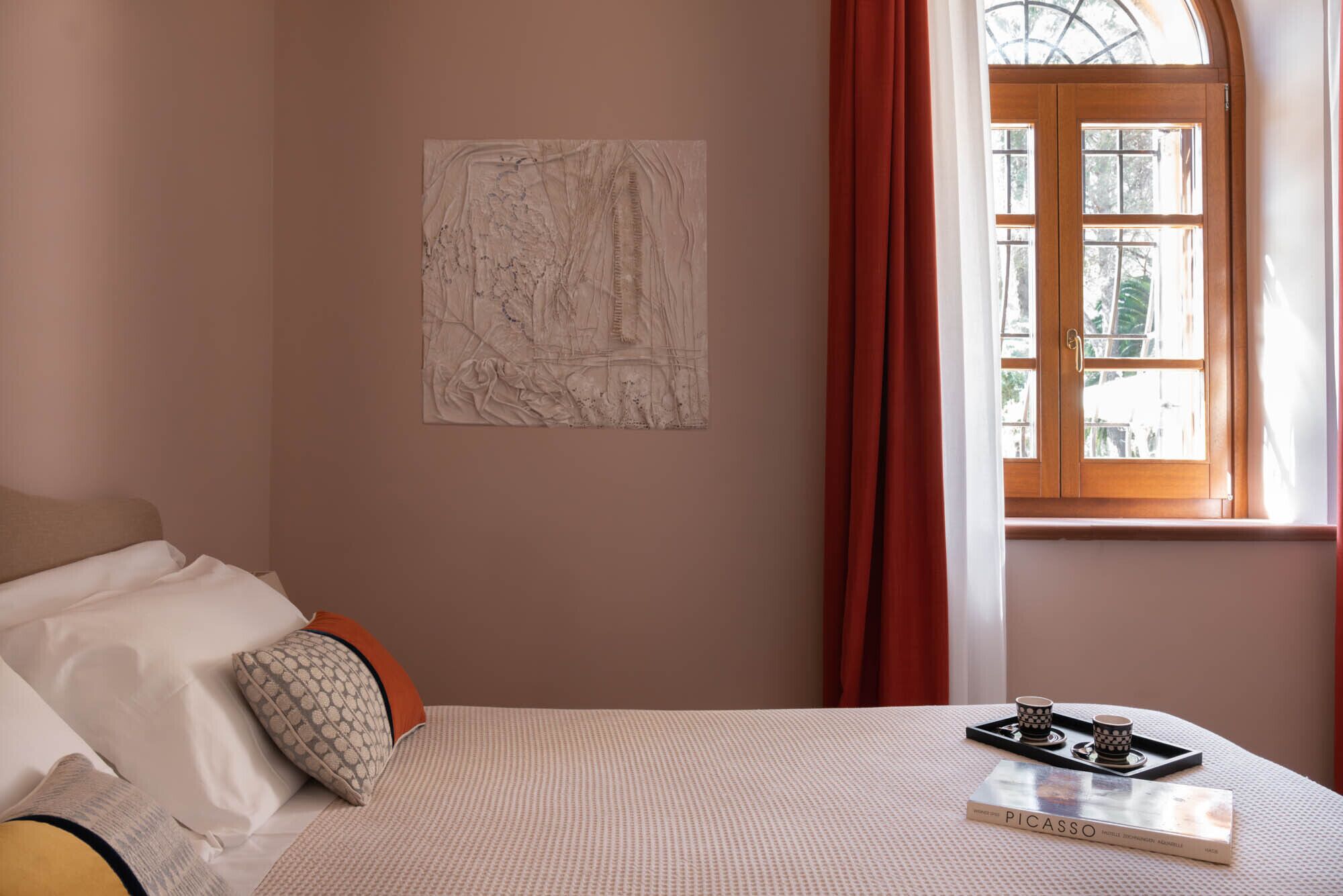 A bedroom with a bed, a window with curtains, a wall-mounted artwork, and a tray with cups and a book.