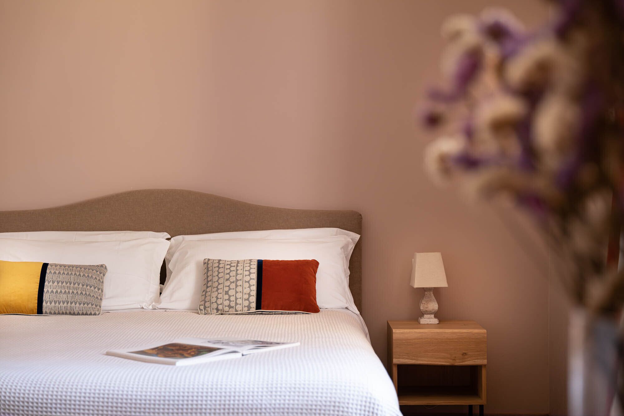 A neatly made bed with white bedding and patterned pillows, a small bedside table with a lamp, and a vase with flowers on the right side.