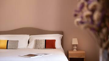 A neatly made bed with white bedding and patterned pillows, a small bedside table with a lamp, and a vase with flowers on the right side.