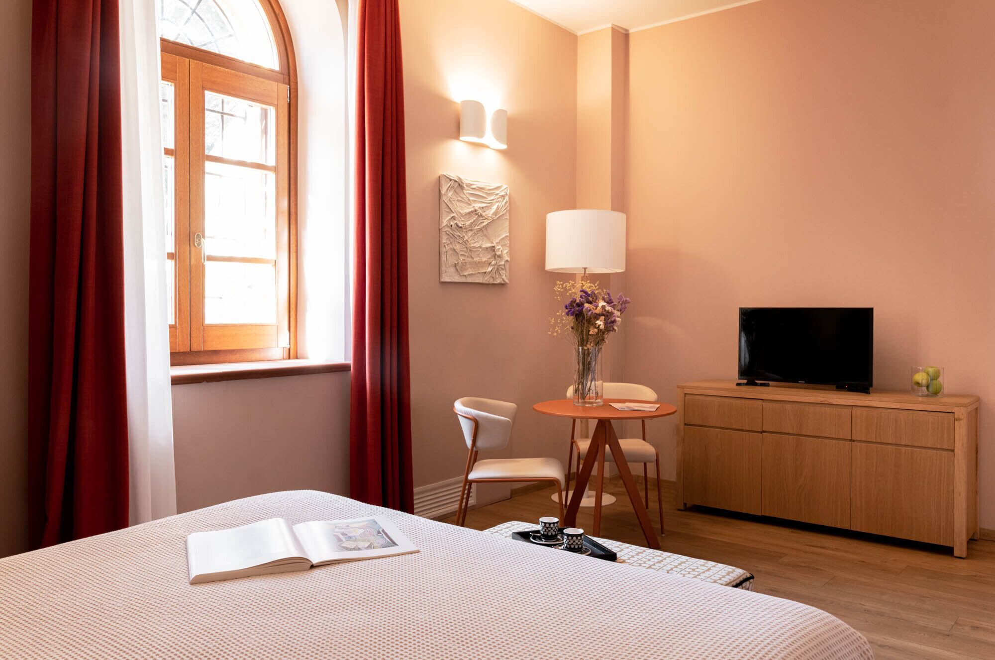 A hotel room with a bed, a desk with a lamp, a television, and a small table with a vase of flowers.