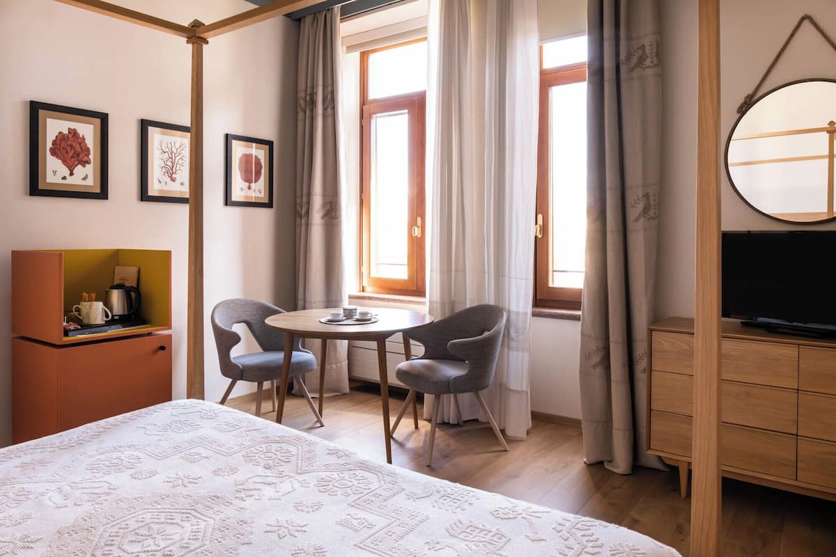 A hotel room with a bed, a small table with chairs, a TV, and framed botanical prints on the wall.