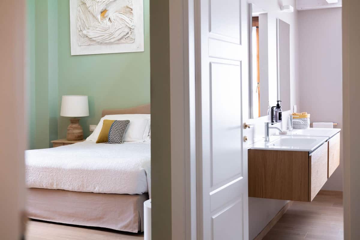 A hotel room with a bed, bedside table, lamp, and a painting on the wall.