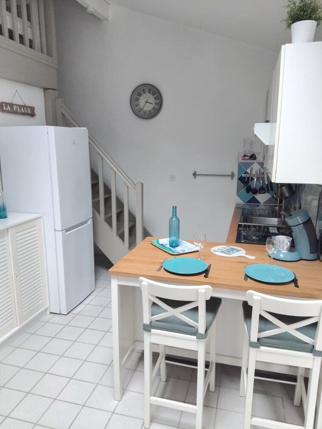 Une cuisine de taille réduite avec une table en bois, des chaises blanches, une horloge et un réfrigérateur.
