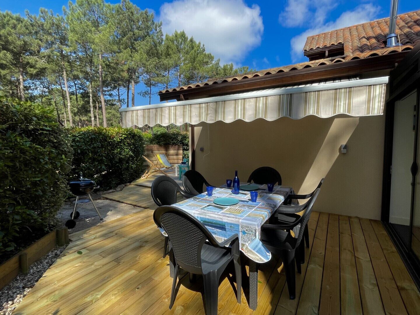 A patio with a table set for two, surrounded by chairs, a grill, and a covered area with a striped awning.