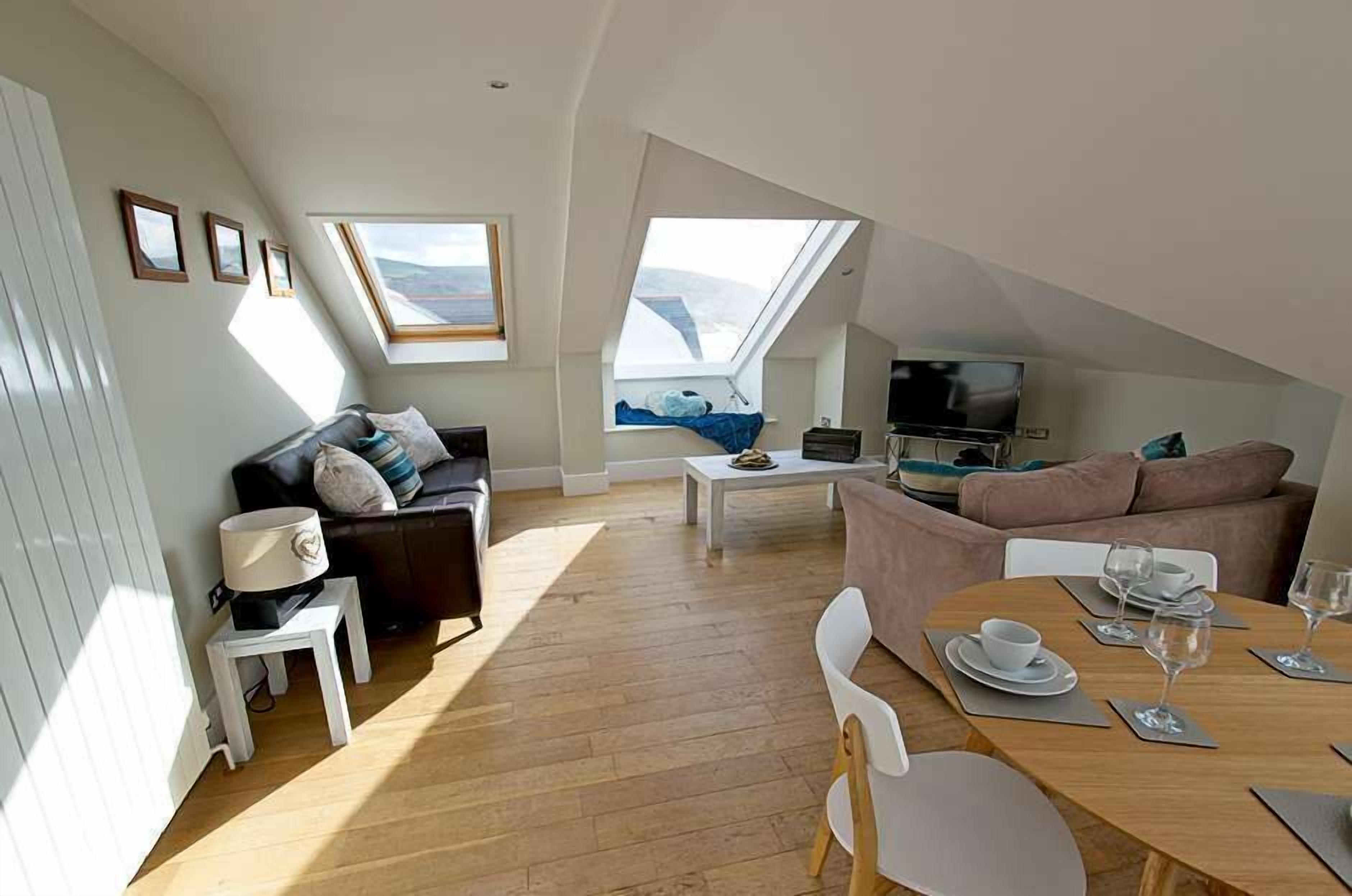 A loft-style living room with a high ceiling, skylights, a sofa, a dining table set, and a television.