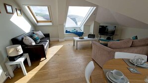 Un appartement moderne de style « loft » avec de hauts plafonds, des fenêtres de toit, un canapé en cuir noir, un canapé marron, une table à manger pour deux personnes et un téléviseur à écran plat.