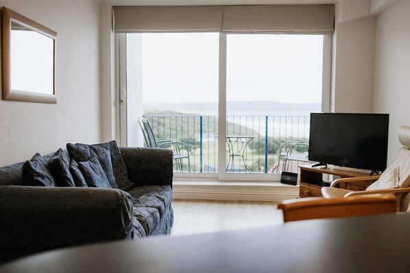 A living room with a sofa, a TV, and a chair. There is a balcony with a view of the landscape.