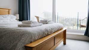 A neatly made bed with folded towels, a wooden headboard, and a view of a balcony through a large window. - Cosy 2 bed apartment close to village with 3 balconies and seaviews! (Woolacombe)